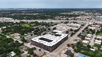 A large building with a flat roof is surrounded by smaller buildings and greenery.